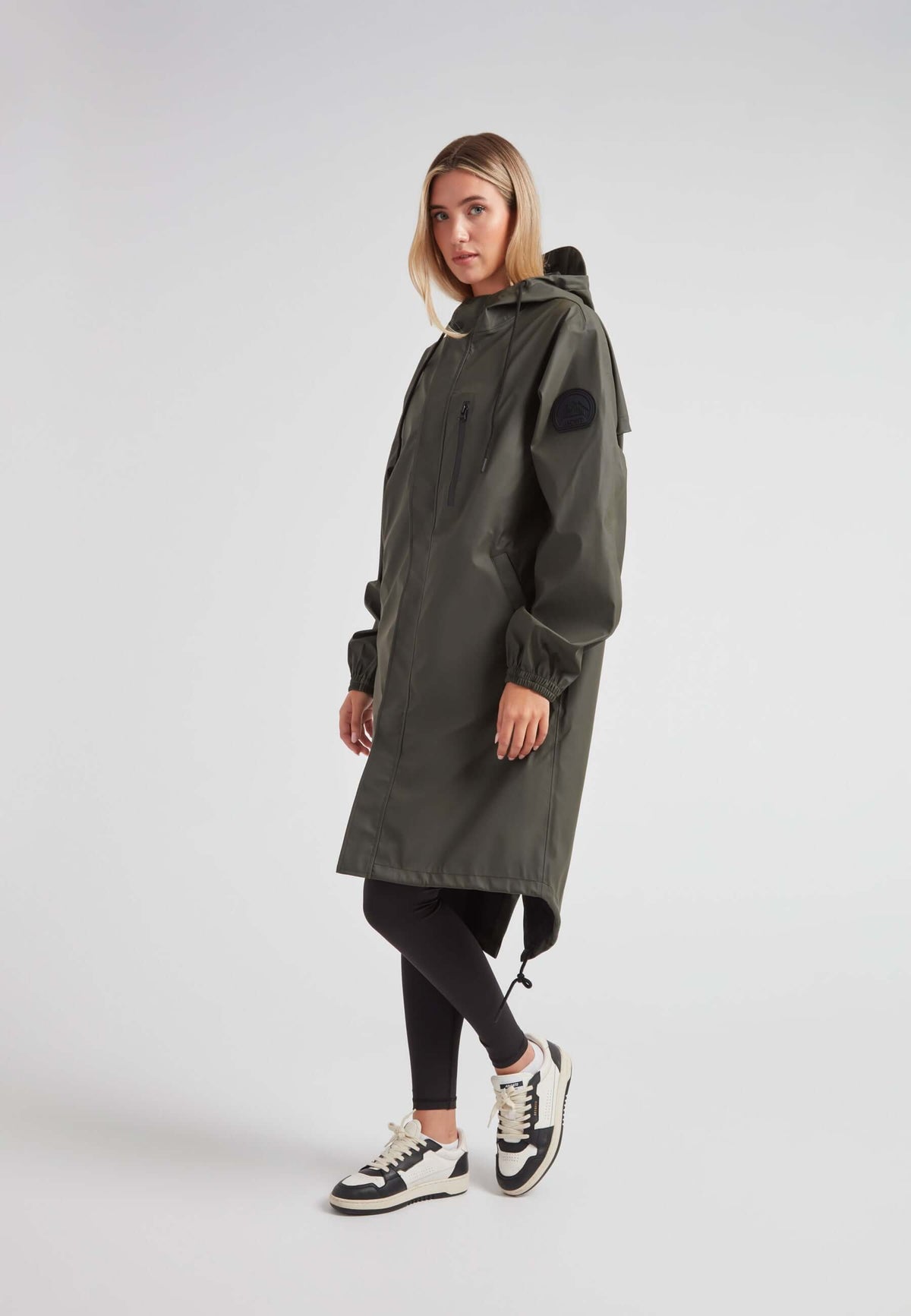 Woman wearing ELEMENTS unisex raincoat in khaki with hood and pockets, styled with leggings and sneakers.