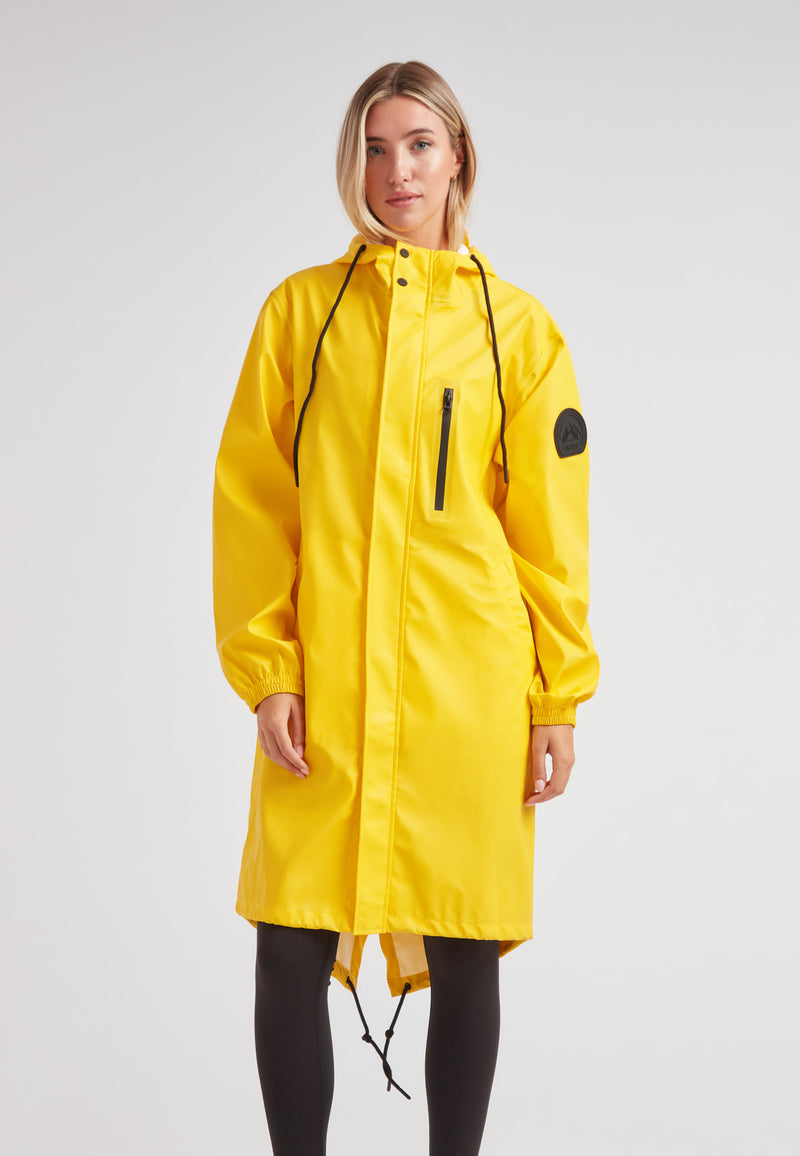 Woman wearing a yellow Elements longline raincoat 2.0 with a zippered pocket and drawstring hood.