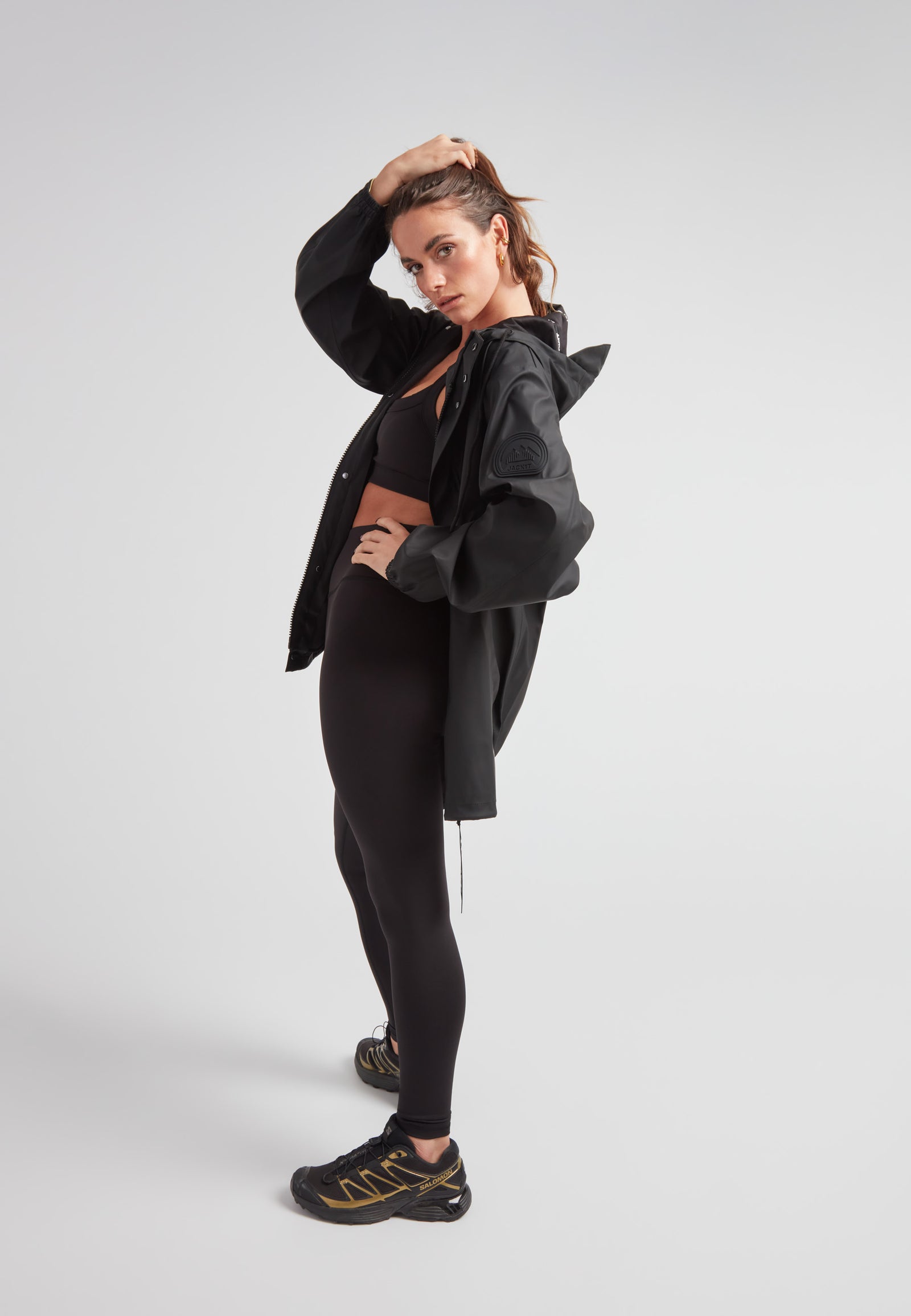 Woman modeling ELEMENTS Raincoat 2.0 Black with black leggings and sneakers in a studio setting.