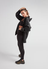 Woman modeling ELEMENTS Raincoat 2.0 Black with black leggings and sneakers in a studio setting.