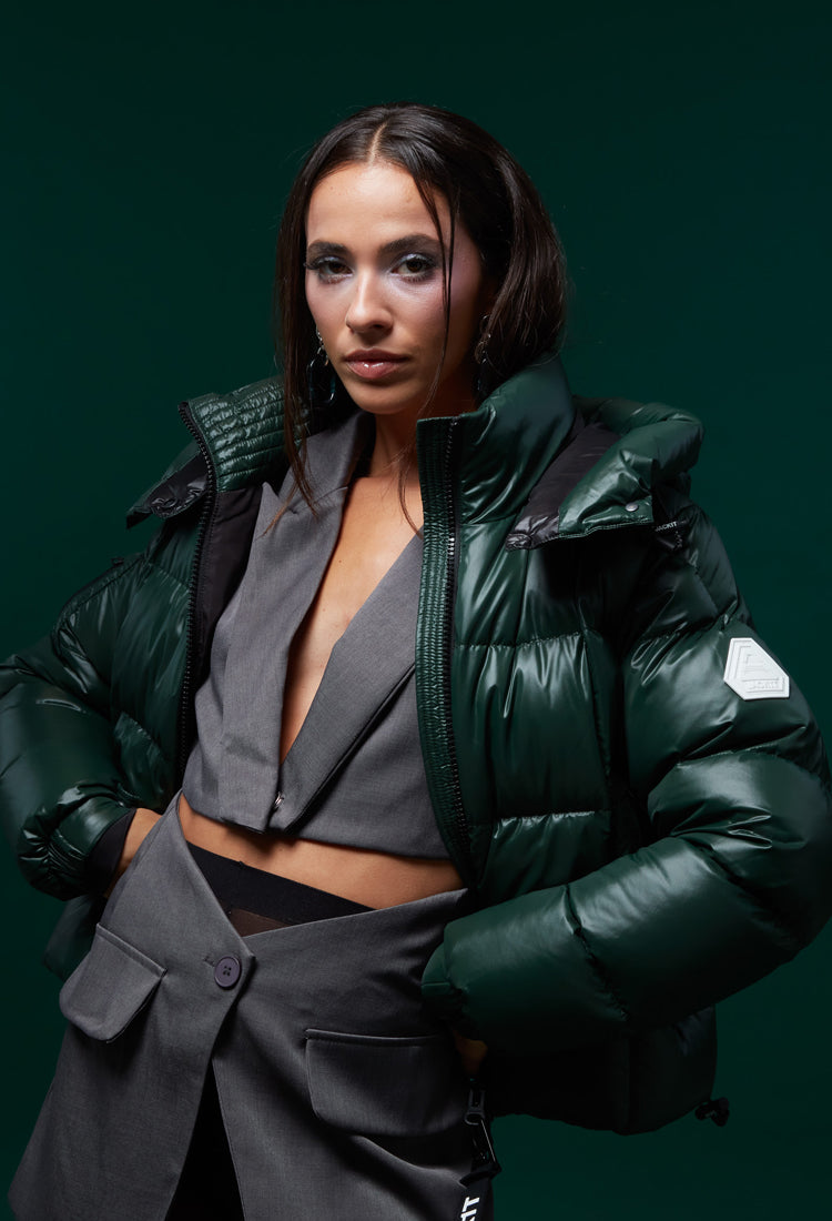 Model wearing a stylish green puffer jacket with a cropped gray top, posing confidently against a dark green background.