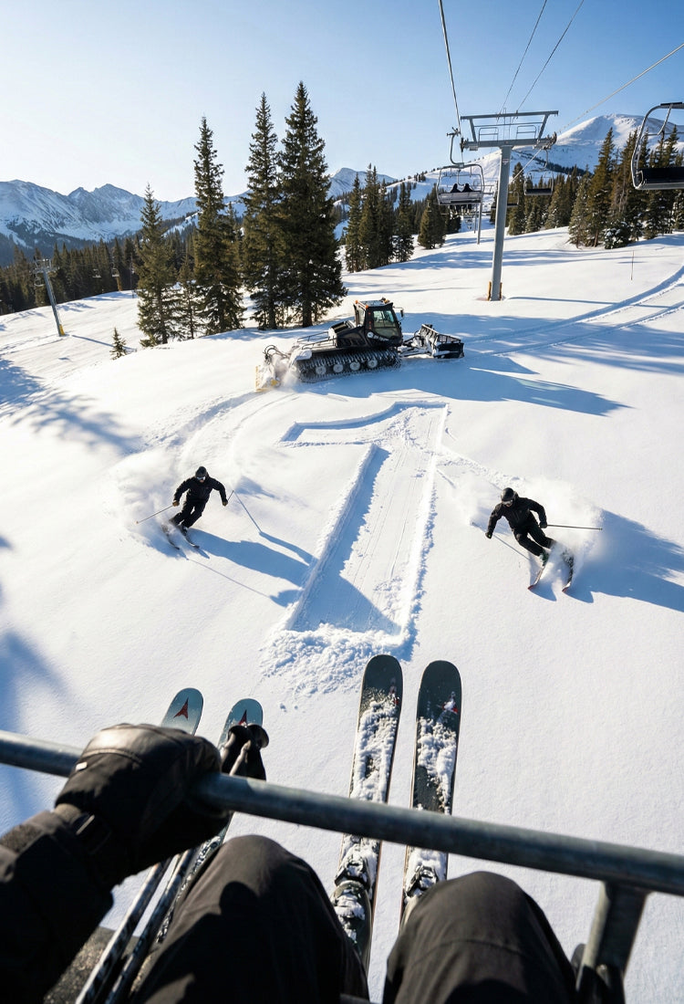 View from a ski lift over skiers carving tracks in fresh snow with a snow groomer in the background.