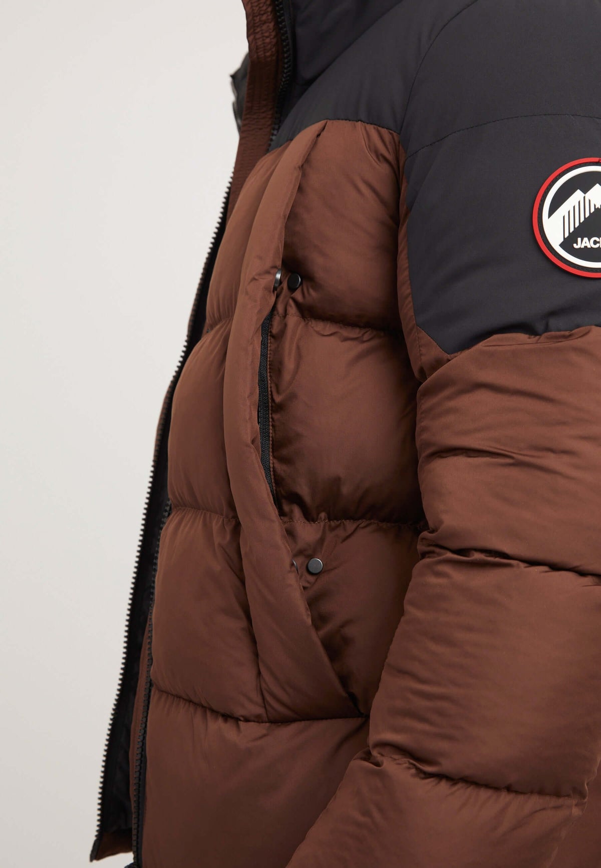 Mens brown puffer jacket showing side view with mid brown and black colors, featuring mid-body pockets and a badge.