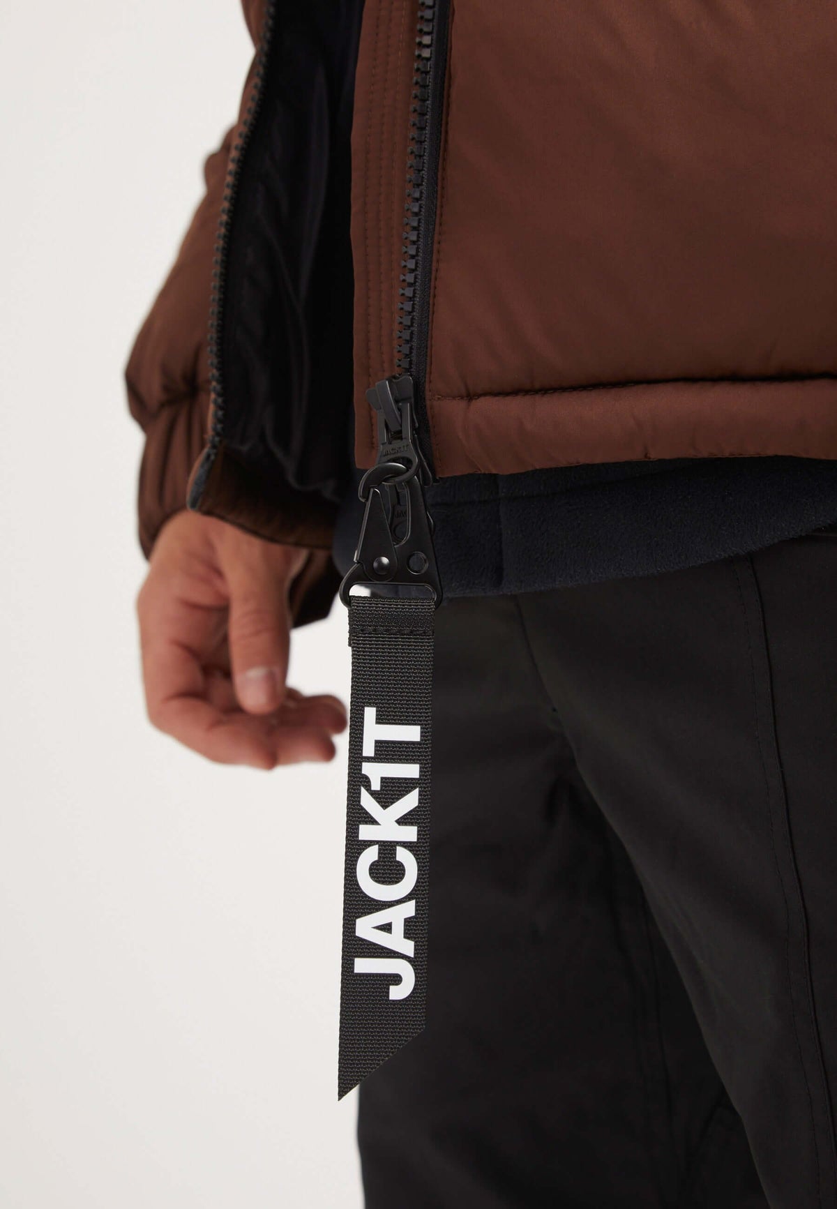 Close-up of a hand holding a zipper on a Luna Puffer Jacket in mid brown color, featuring a JACKIT tag, showcasing the mens brown puffer jacket.