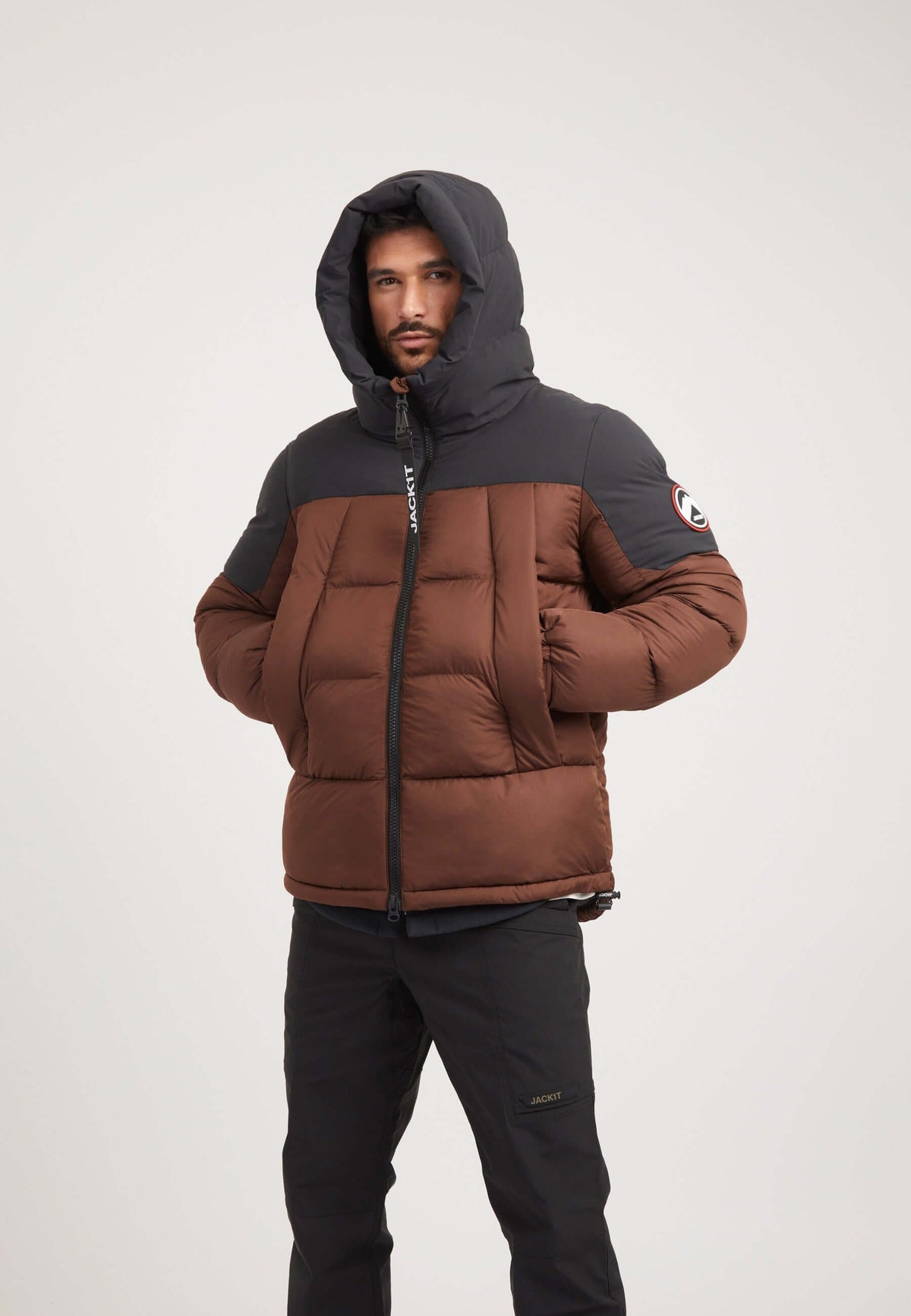 Model wearing a Luna Puffer Jacket in mid brown and black, showcasing a mens brown puffer jacket style with a dropped hem and hood.