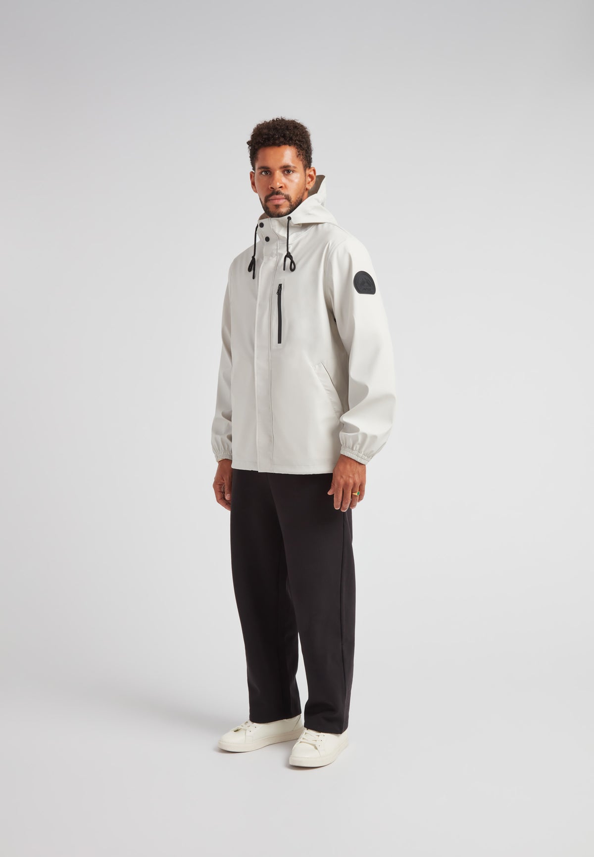 Man wearing ELEMENTS Unisex Raincoat 2.0 Stone with hood and black accents, styled with black pants and white shoes.