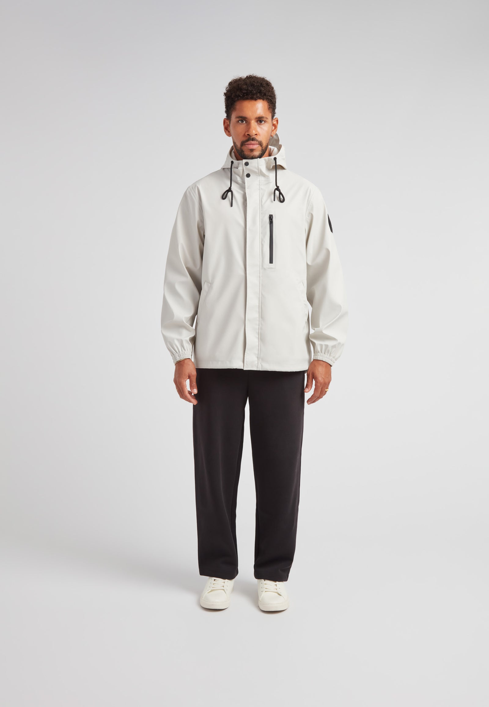 Man wearing ELEMENTS Unisex Raincoat 2.0 Stone with hood and drawstrings, paired with black pants.