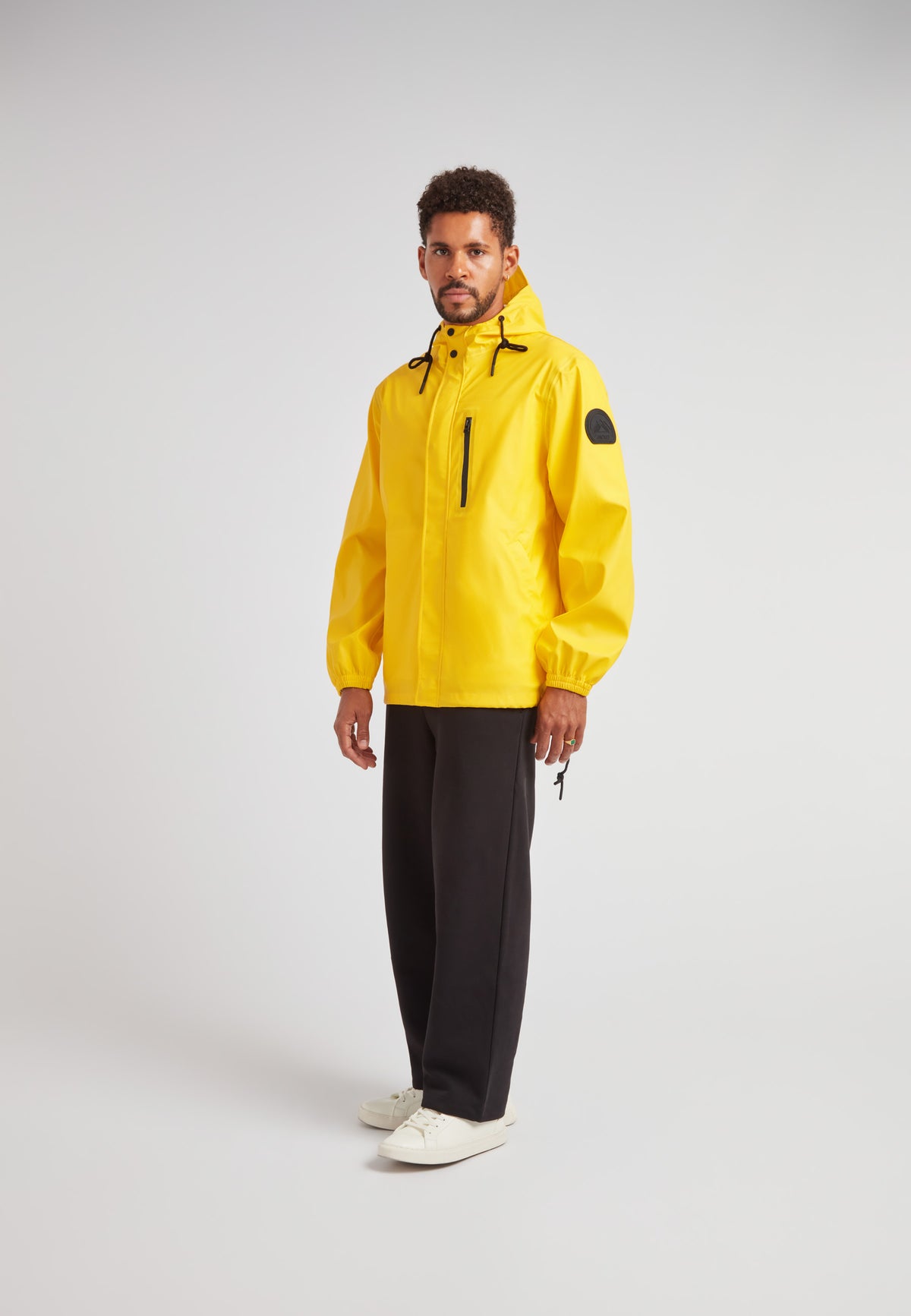 Man wearing ELEMENTS Unisex Raincoat 2.0 Yellow, standing in studio with black pants and white sneakers.