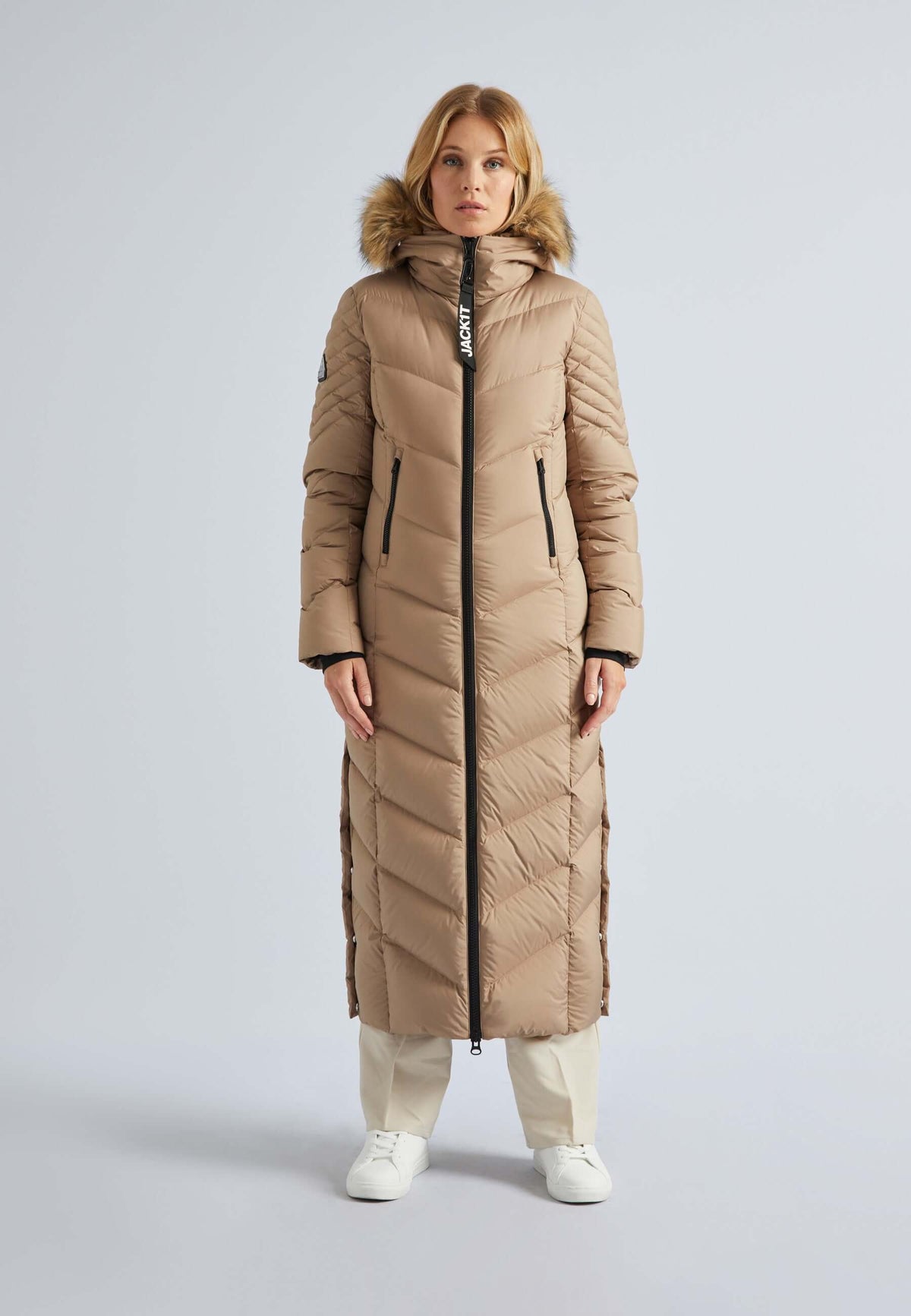 Model wearing an extra long puffer coat womens in beige with fur hood and zippers.