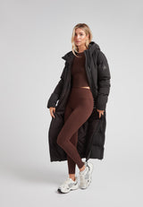 Woman wearing a Black Star Extra Long Down Coat Matt Black over brown legging and top, showcasing its stylish design.