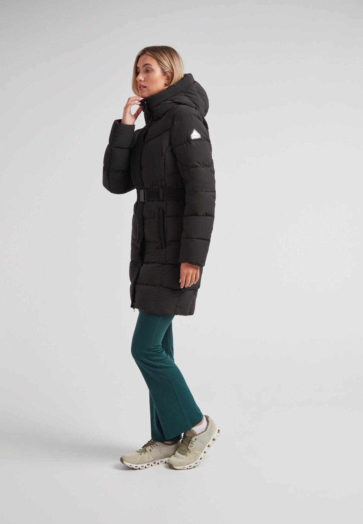 Woman wearing the Diamond Belted Down Coat Matt Black with hood and belt, paired with green leggings and sporty shoes.