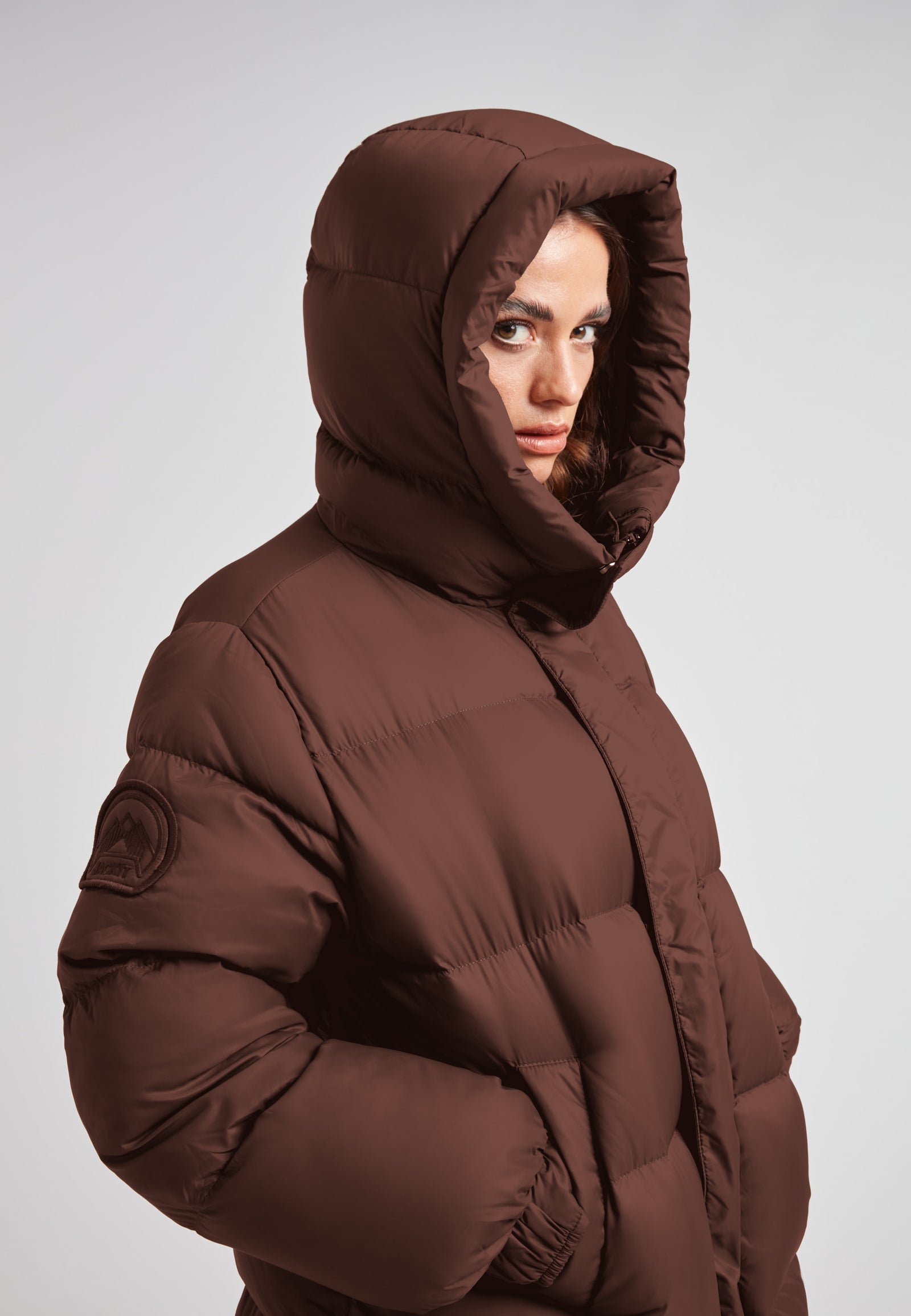 Cocoon Ultra Long Down Coat Mid Brown on model with hood, showcasing its oversized silhouette and warm design.