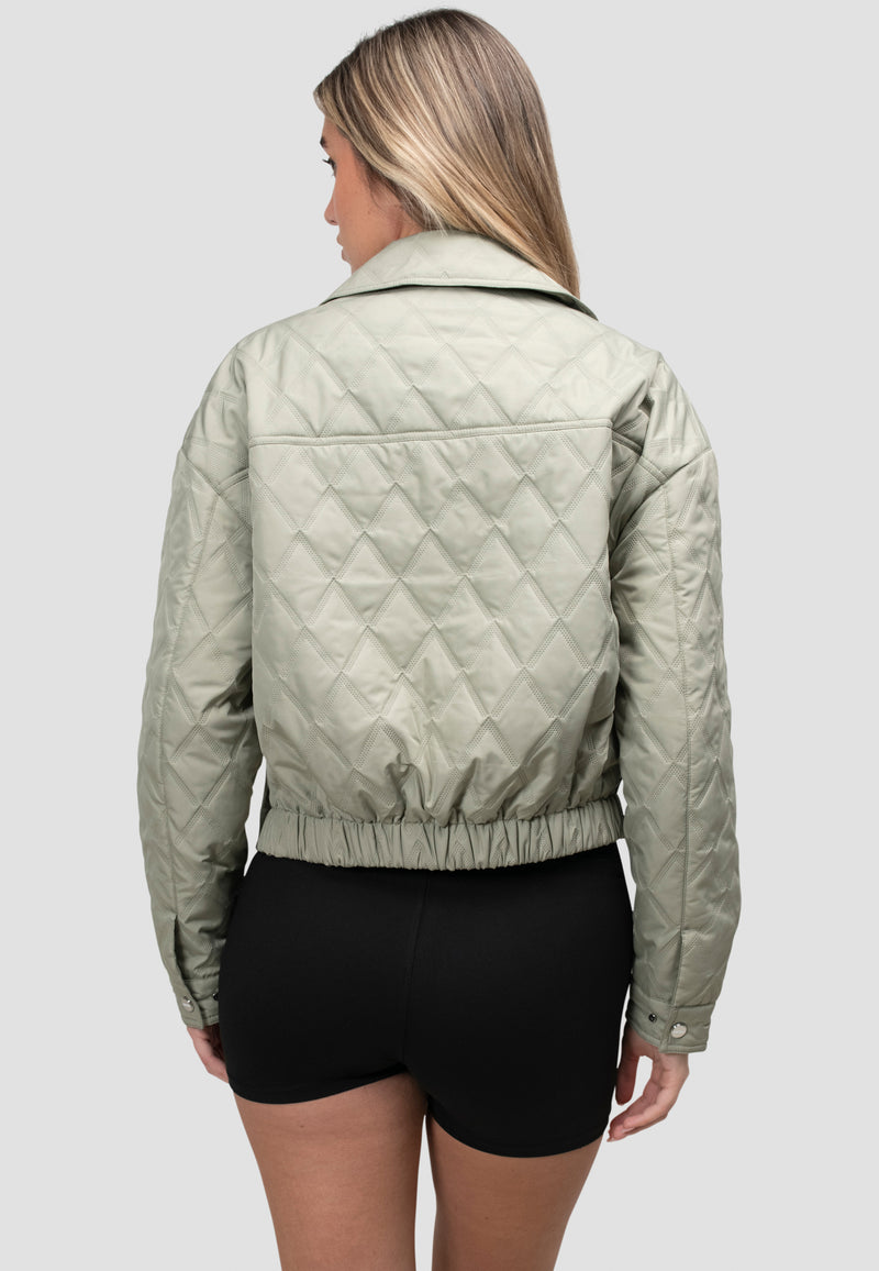 Back view of a woman wearing a women's sage quilted shirt jacket styled with black shorts.