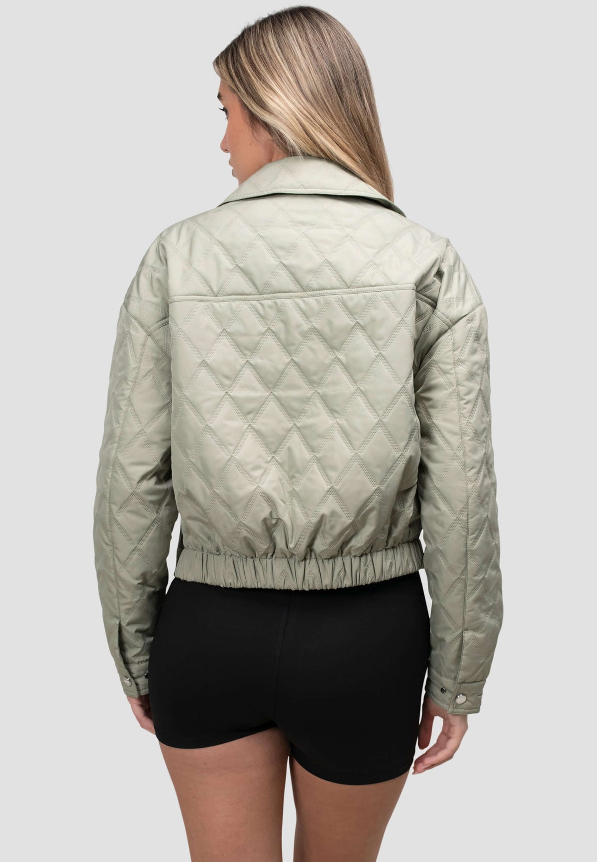 Back view of a woman wearing a women's sage quilted shirt jacket styled with black shorts.