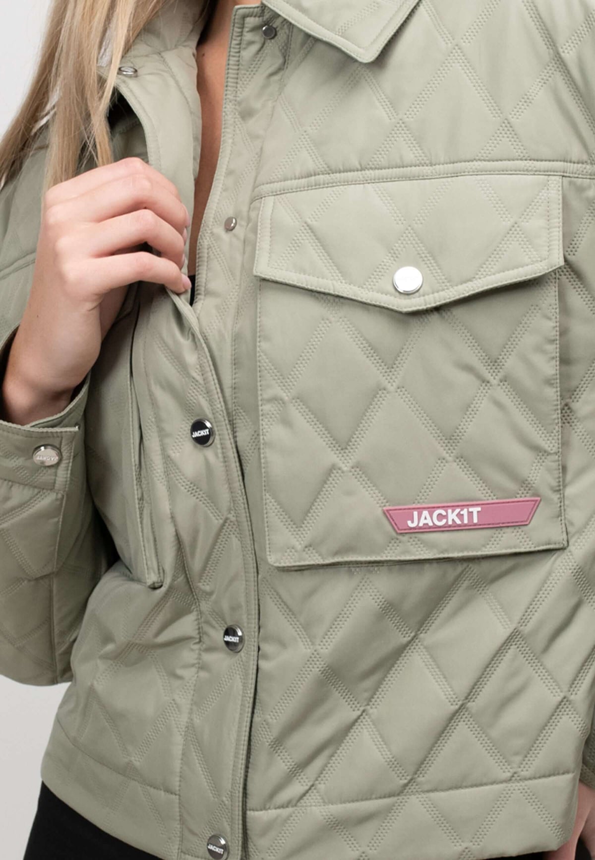 Close-up of a women's sage quilted shirt jacket featuring a buttoned pocket and brand logo detail.