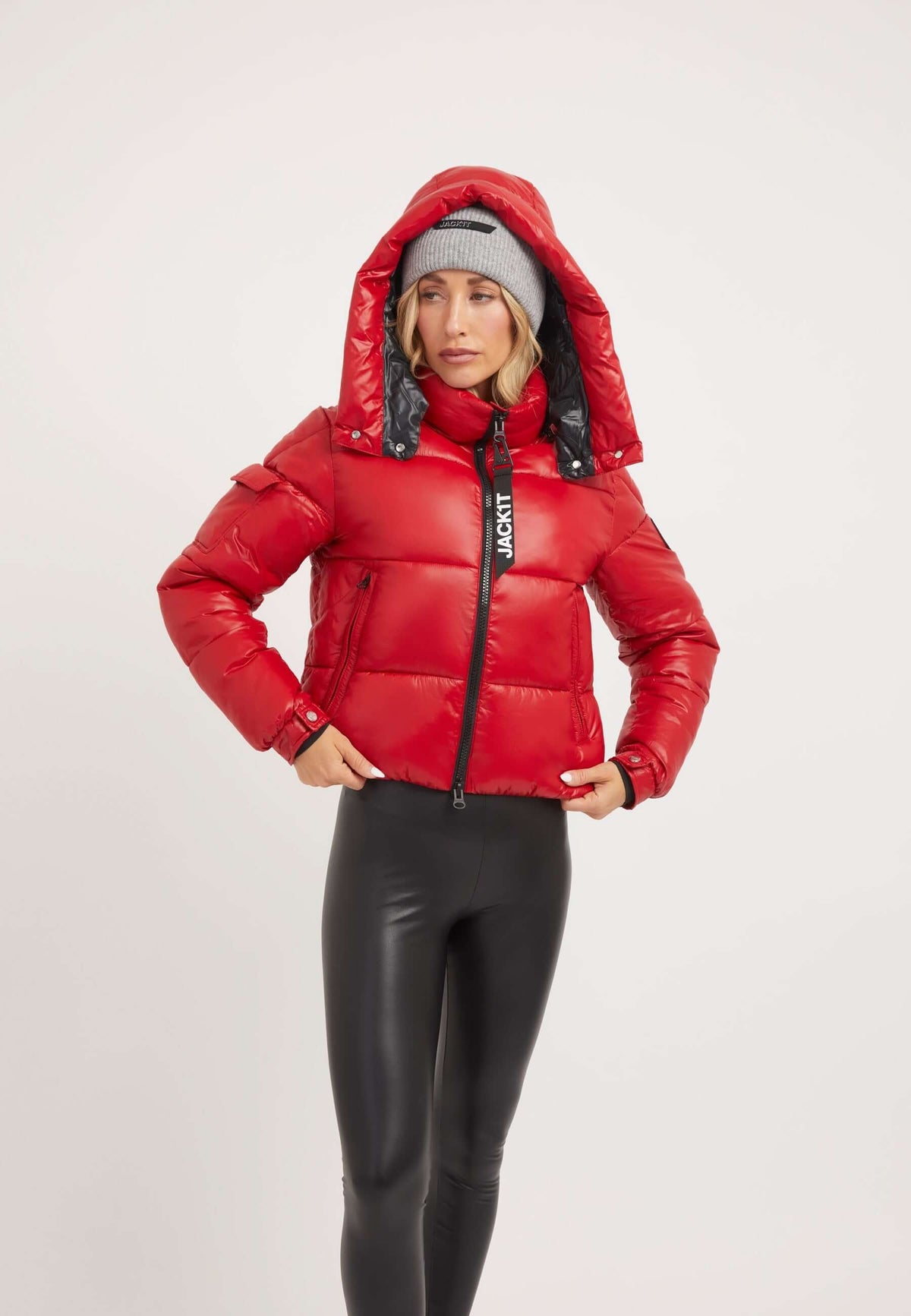 Model wearing a red puffer jacket with a removable hood and ski straps, styled with a gray beanie and black leggings.
