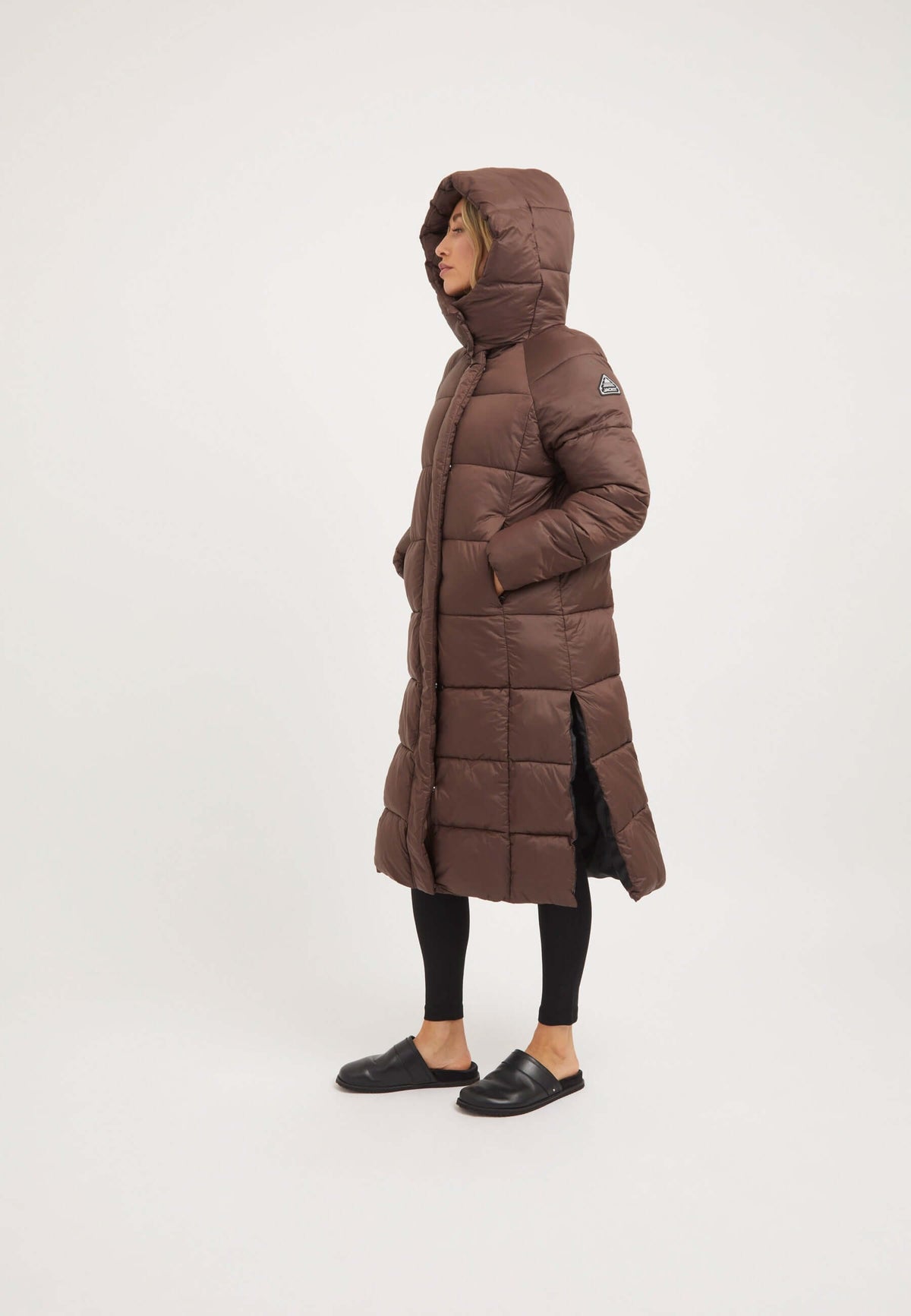 Woman wearing a mid brown womens long puffer coat with a removable hood and side split opening.