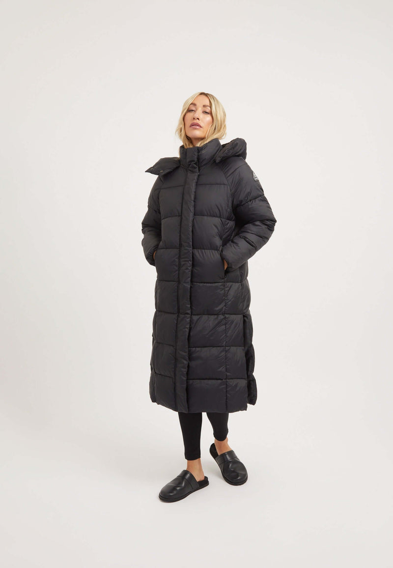 Model wearing the Cloud Boss Coat Black, a womens long puffer coat, featuring a full length silhouette and removable hood.