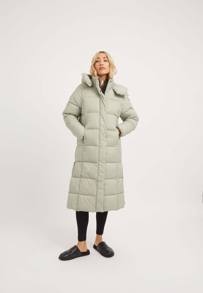 Model wearing the Cloud Boss Coat Sage, a womens long puffer coat with a full length silhouette and removable hood.