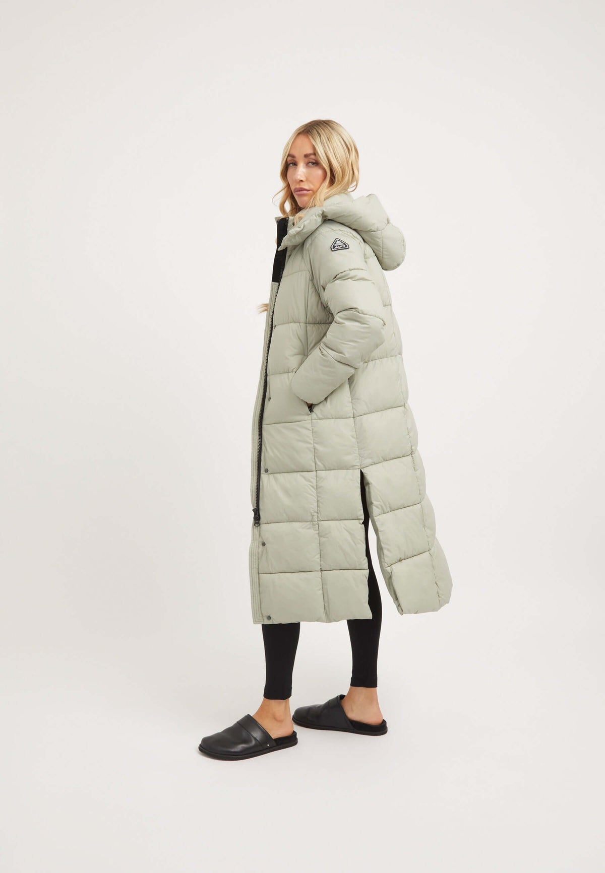 Model wearing the Cloud Boss Coat Sage, a womens long puffer coat with a full length silhouette and removable hood.