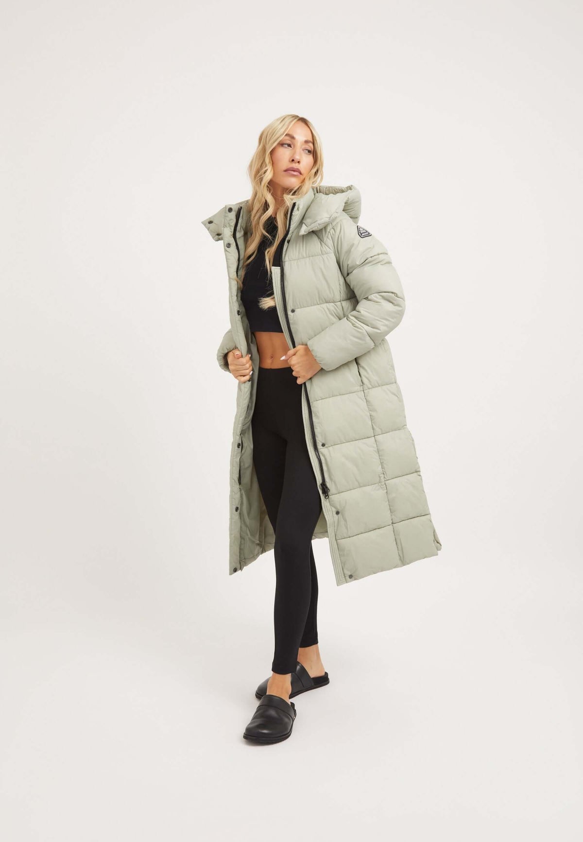 Model wearing a Cloud Boss Coat Sage, a womens long puffer coat, featuring a full length silhouette and removable hood.