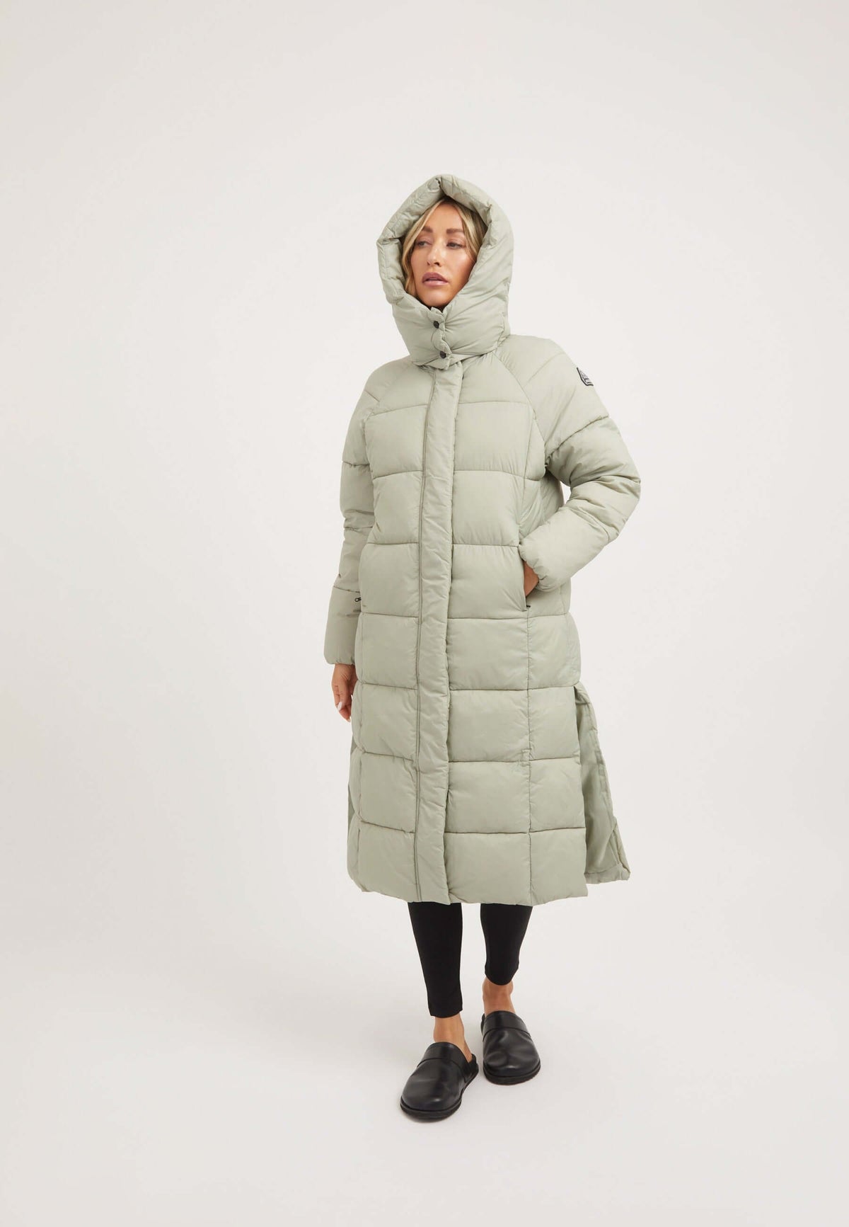 Model wearing a Cloud Boss Coat Sage, a womens long puffer coat with a full length silhouette and removable hood.
