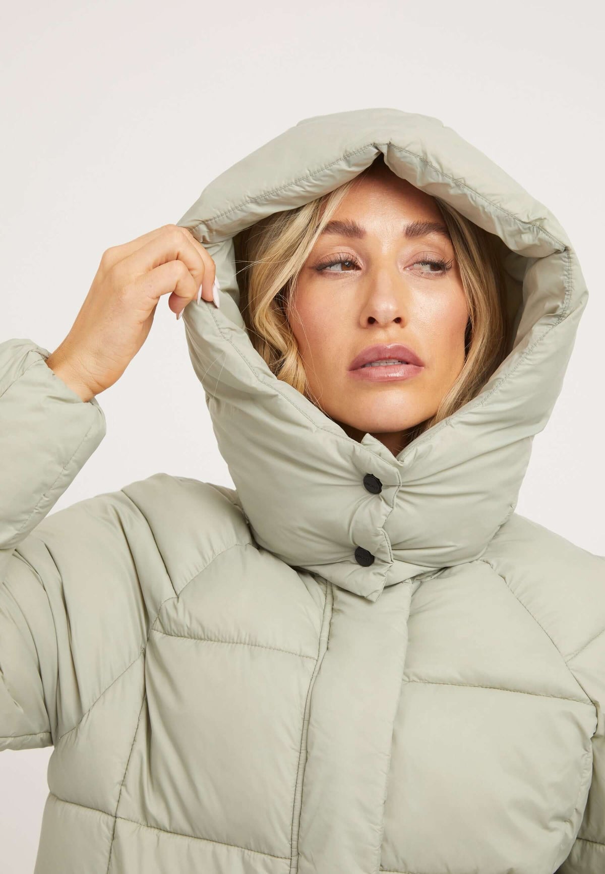 Close-up of a woman wearing a Cloud Boss Coat Sage with a removable hood, showcasing the stylish design of the womens long puffer coat.