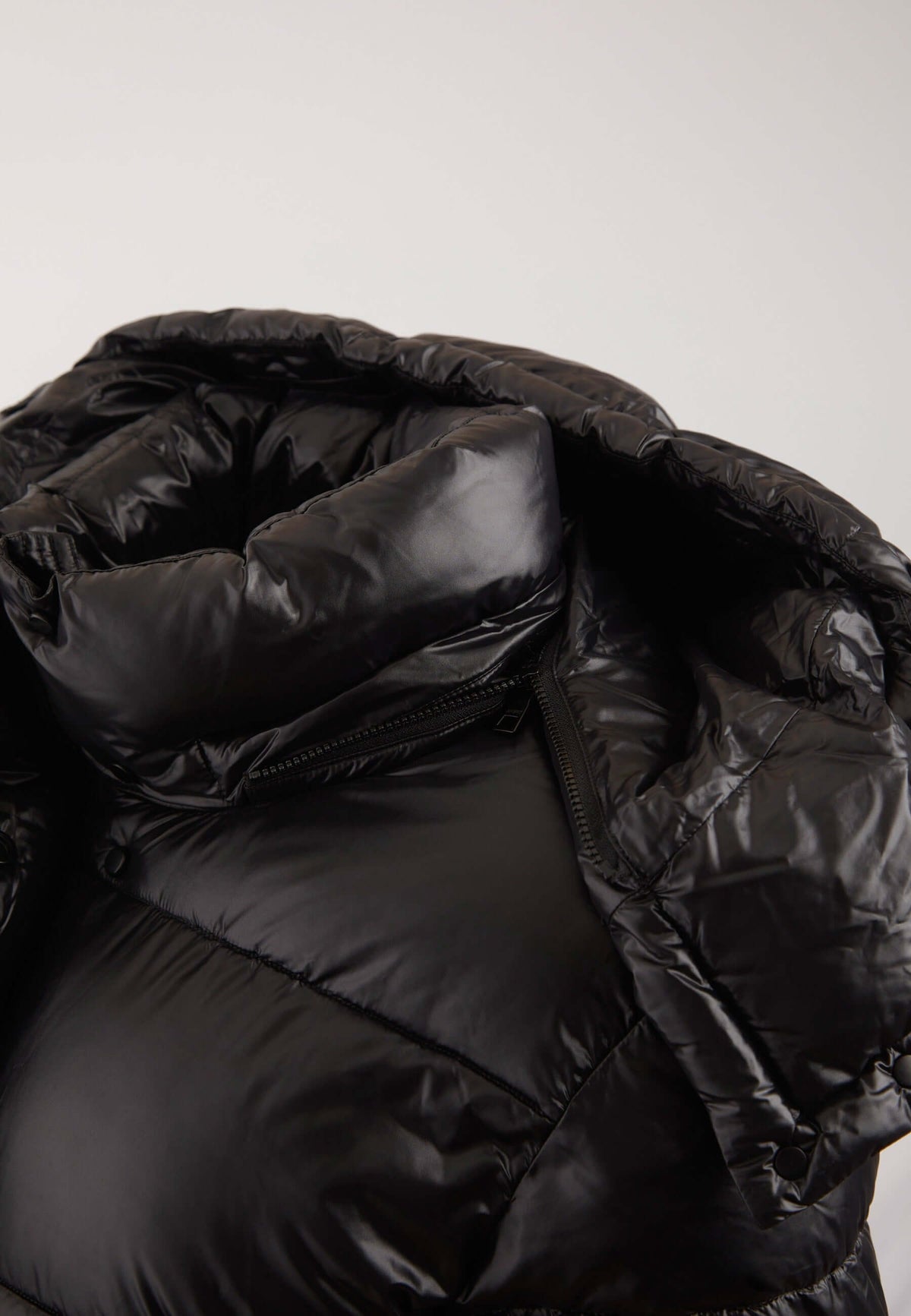Close-up of a black puffer jacket with quilted padding and a glossy finish, showing the hood and zipper details against a light gray background.