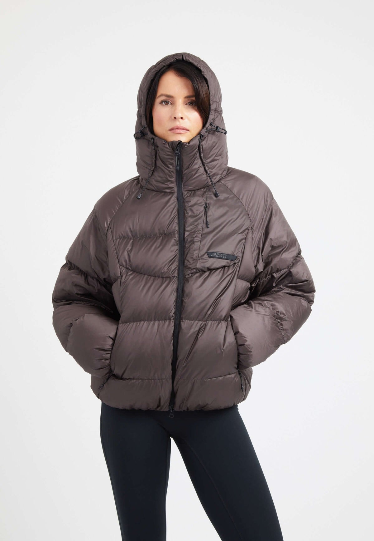 Woman wearing an oversized puffer brown jacket with a hood and zip pockets, showcasing its modern silhouette and down fill.