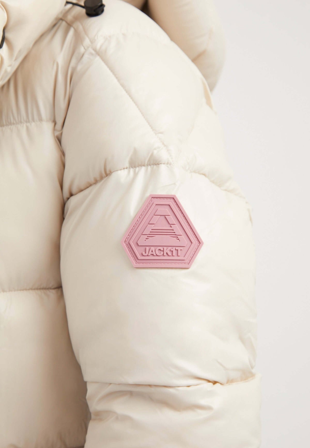 Close-up of the sleeve of a womens belted puffer coat featuring a logo patch in pink on a cream-colored puffer