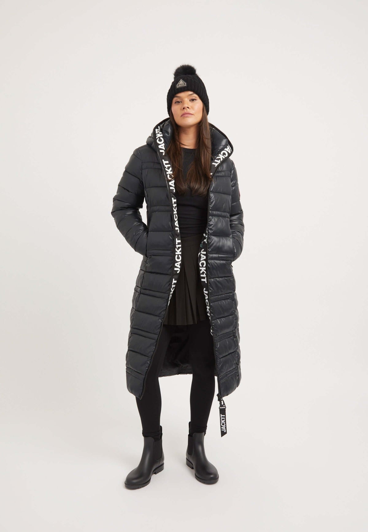 Woman wearing a black R4D Extra Long Puffer Coat with a hood, styled with a skirt and beanie, showcasing warmth and fashion.