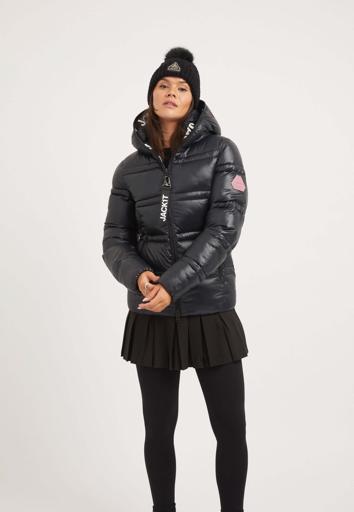 Model wearing a ladies black puffer jacket with a hood, paired with a pleated skirt and beanie.