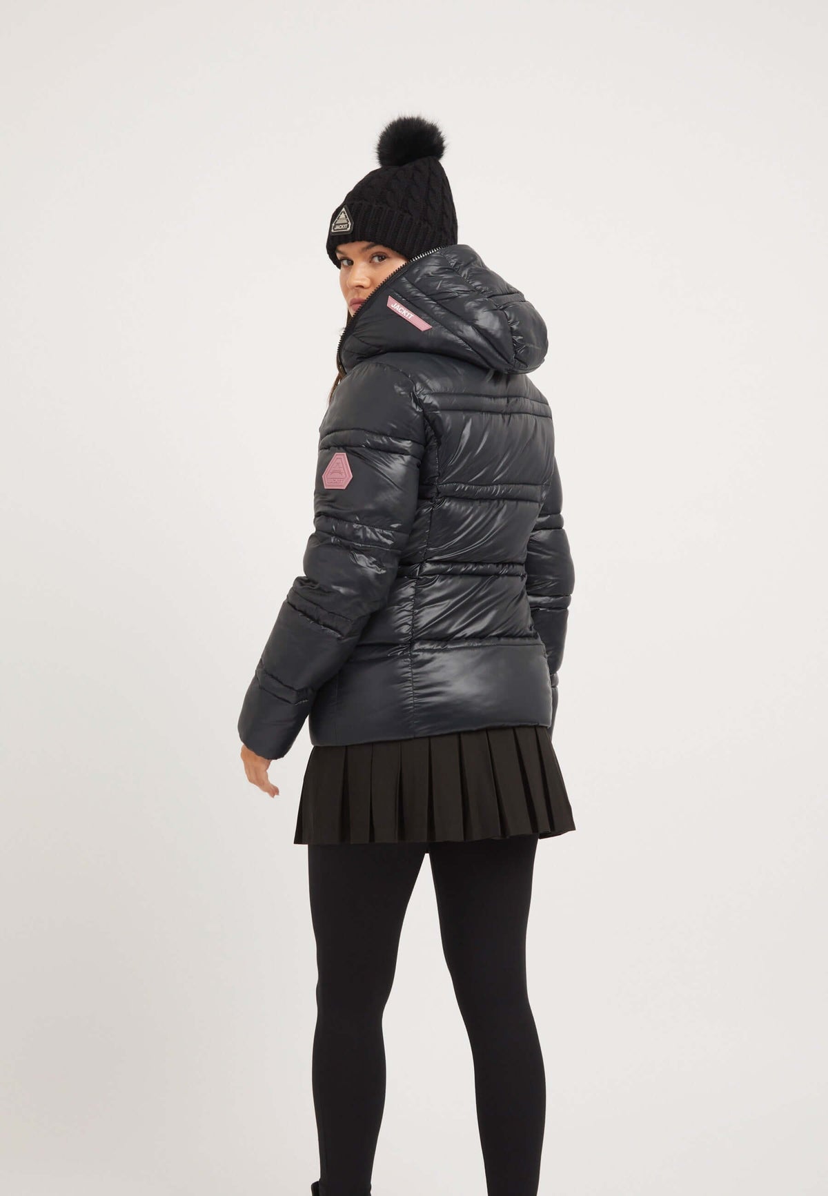 Back view of a model wearing a ladies black puffer jacket with a hood, styled with a pleated skirt and beanie.