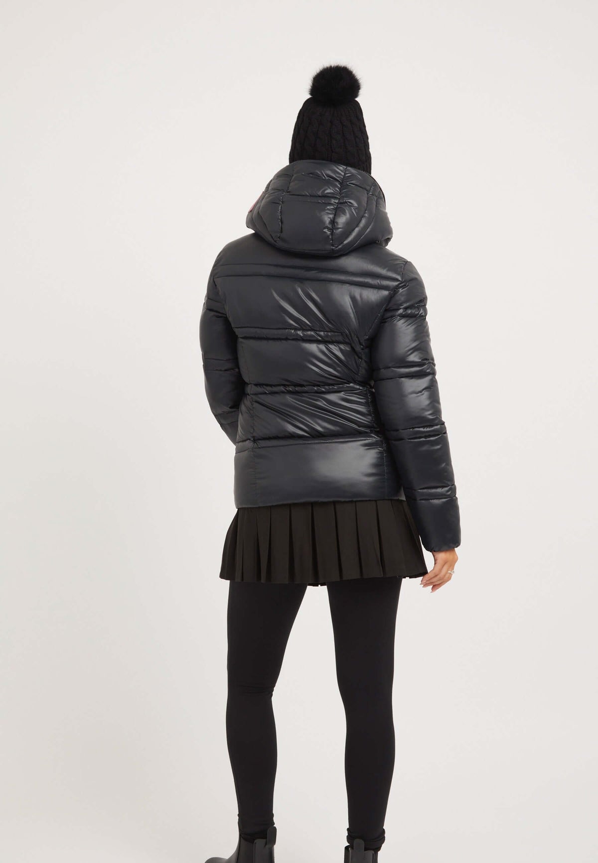 Back view of a model wearing a ladies black puffer jacket with a pleated skirt and knitted hat.
