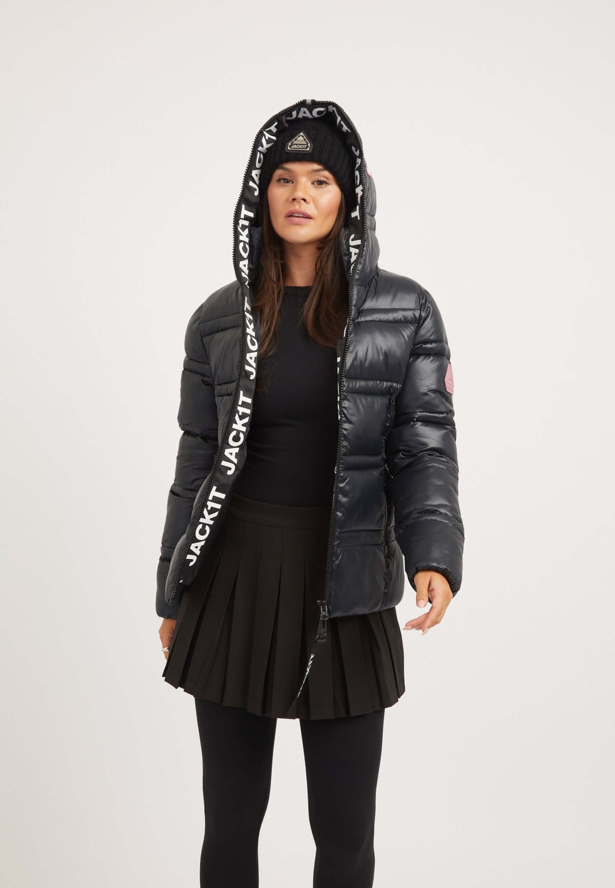 A model wearing a ladies black puffer jacket, featuring a hood, stylish design, and a pleated skirt.