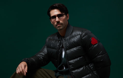 Stylish man wearing a black puffer jacket and sunglasses against a dark green background.