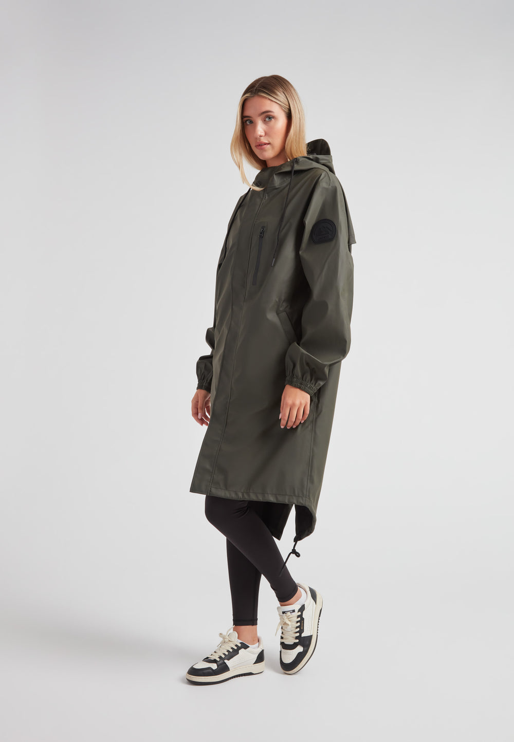 Woman wearing ELEMENTS unisex raincoat in khaki with hood and pockets, styled with leggings and sneakers.