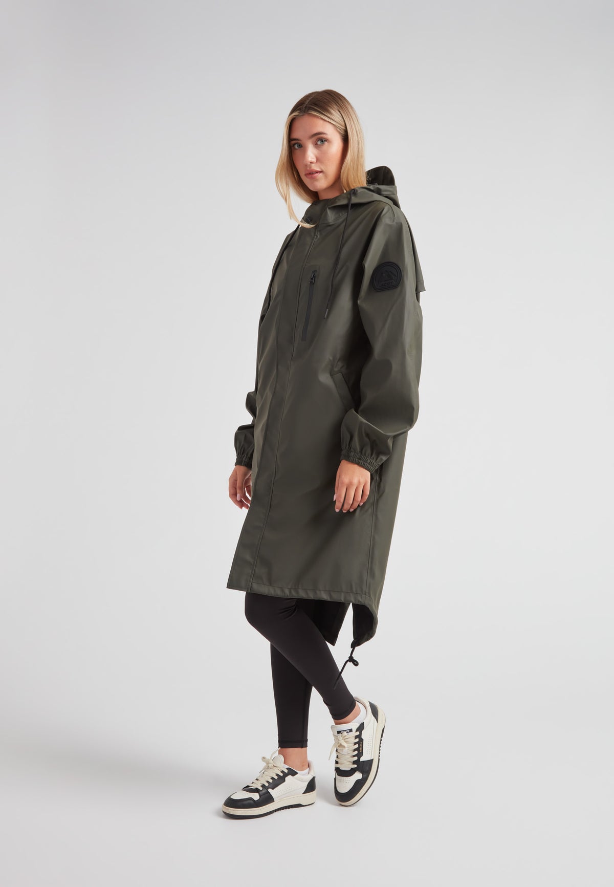 Woman wearing ELEMENTS unisex raincoat in khaki with hood and pockets, styled with leggings and sneakers.