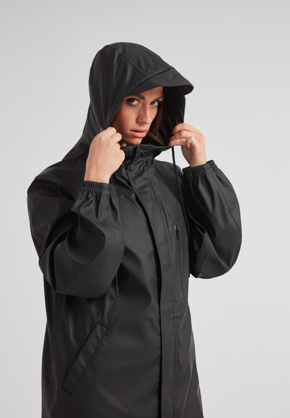 Woman adjusting hood on the ELEMENTS Raincoat 2.0 Black, a stylish and versatile waterproof jacket.