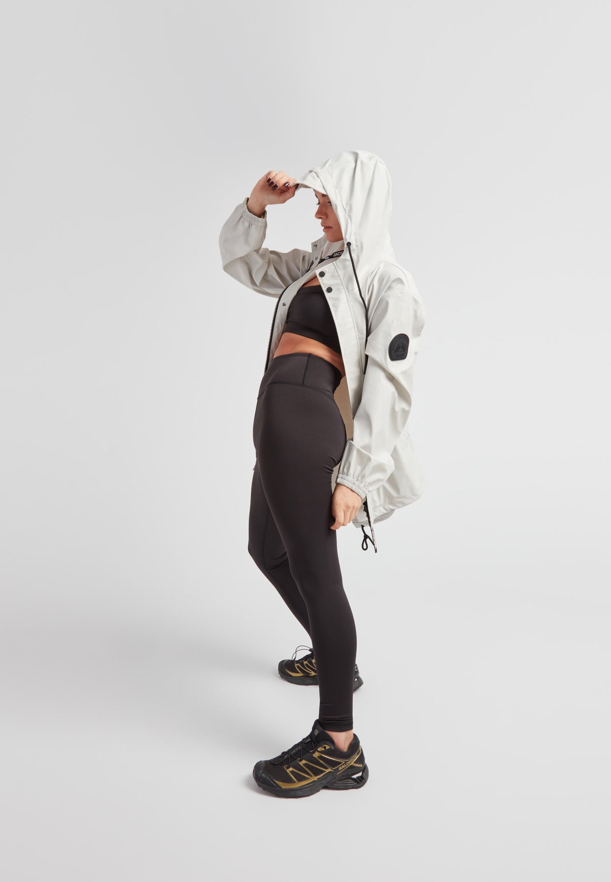 A person wearing the ELEMENTS Raincoat 2.0 Stone with a hood, paired with black leggings and sneakers.