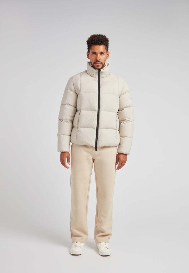 Man wearing the Cloud Lightweight Puffer Racer Jacket Stone with light beige puffer jacket and cream pants.