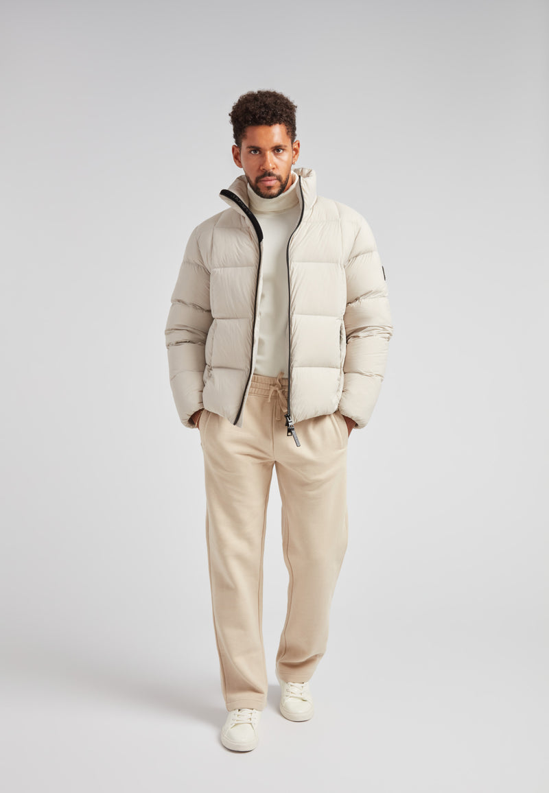 Model wearing Cloud Lightweight Puffer Racer Jacket Stone with casual pants on gray background.