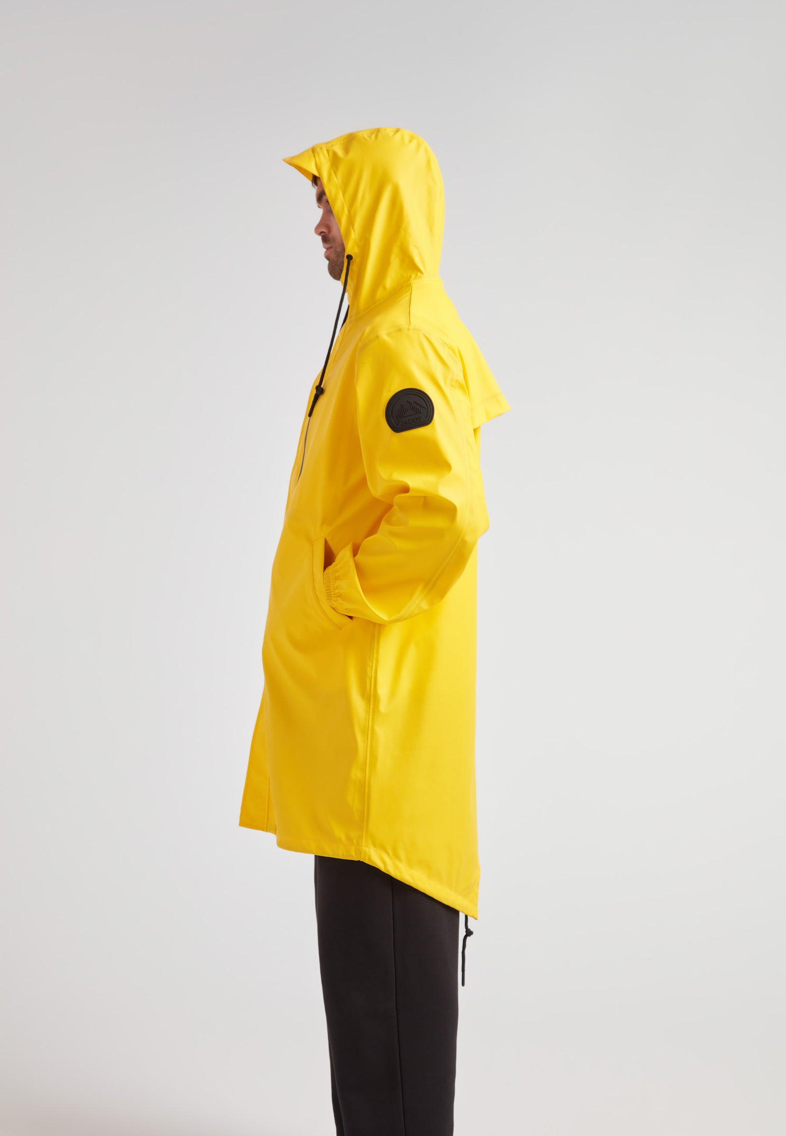 Side view of a person wearing the yellow ELEMENTS longline raincoat 2.0, featuring a hood and pockets.