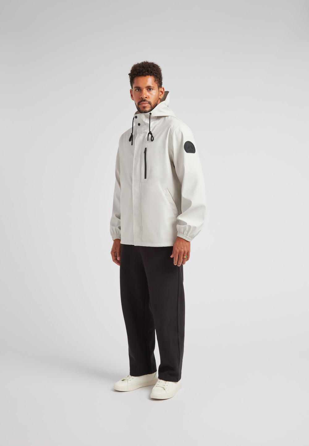 Man wearing ELEMENTS Unisex Raincoat 2.0 Stone with hood and black accents, styled with black pants and white shoes.
