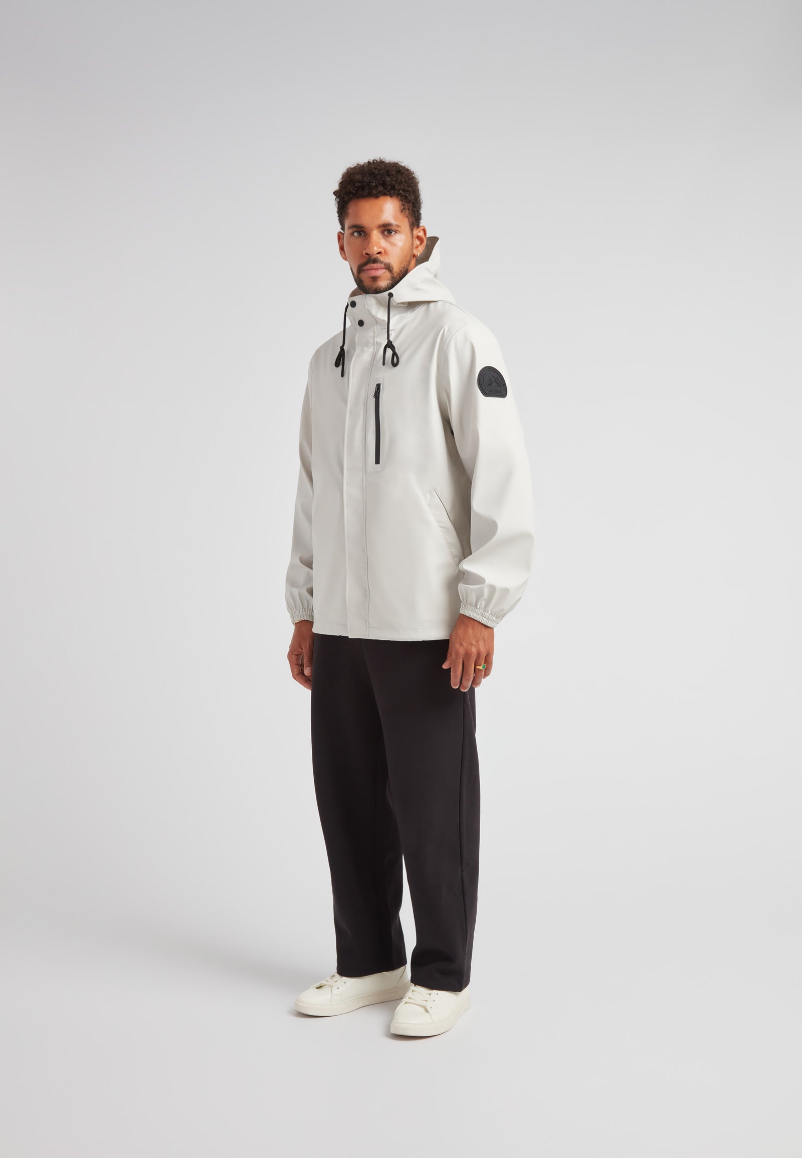 Man wearing ELEMENTS Unisex Raincoat 2.0 Stone with hood and black accents, styled with black pants and white shoes.