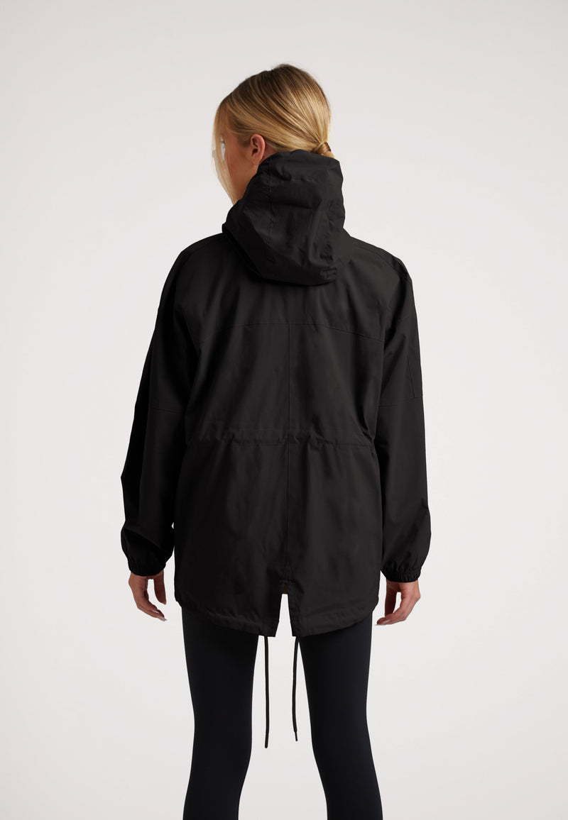 Back view of model wearing the ISOTOPE Ladies lightweight black windbreaker with hood and adjustable hem.
