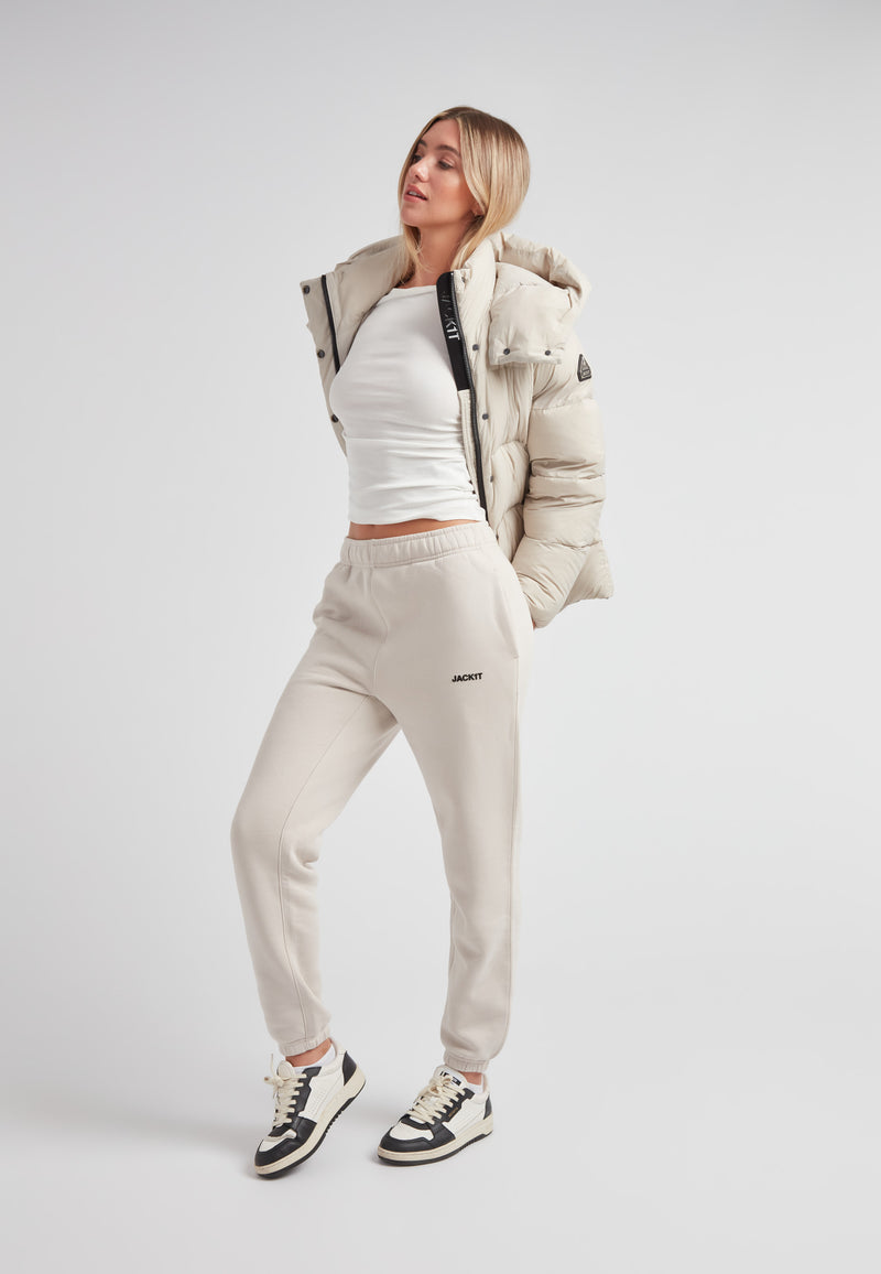 Woman wearing Cloud Prime Longline Down Puffer Jacket Stone with white top and sweatpants, posing stylishly.
