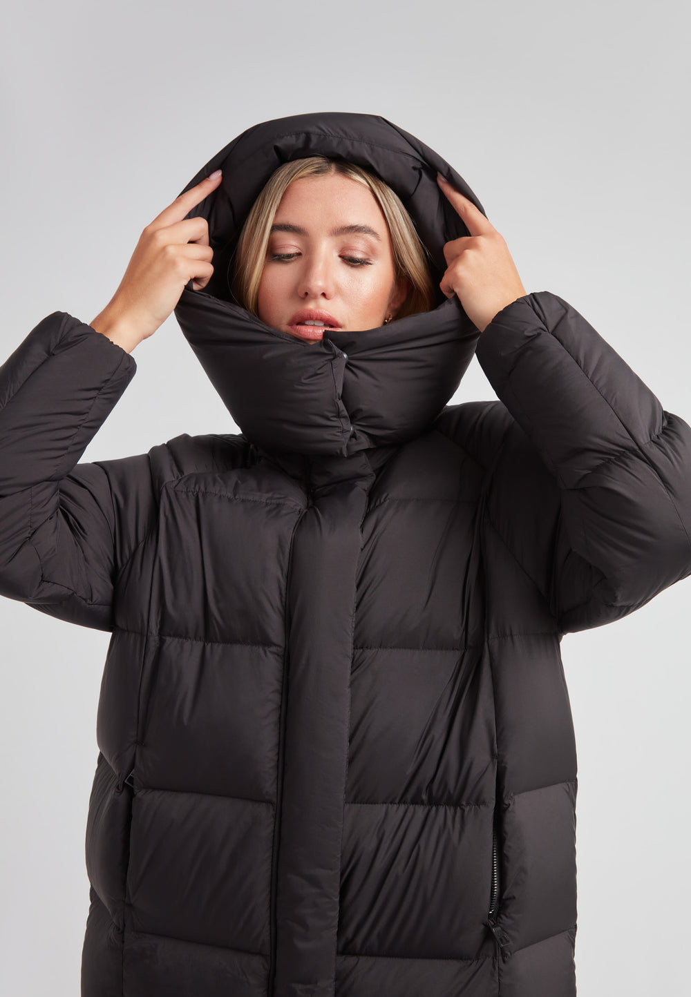 Woman adjusting hood on Cloud Ultra Long Down Puffer Coat, the ideal long puffer jacket in black for winter warmth.