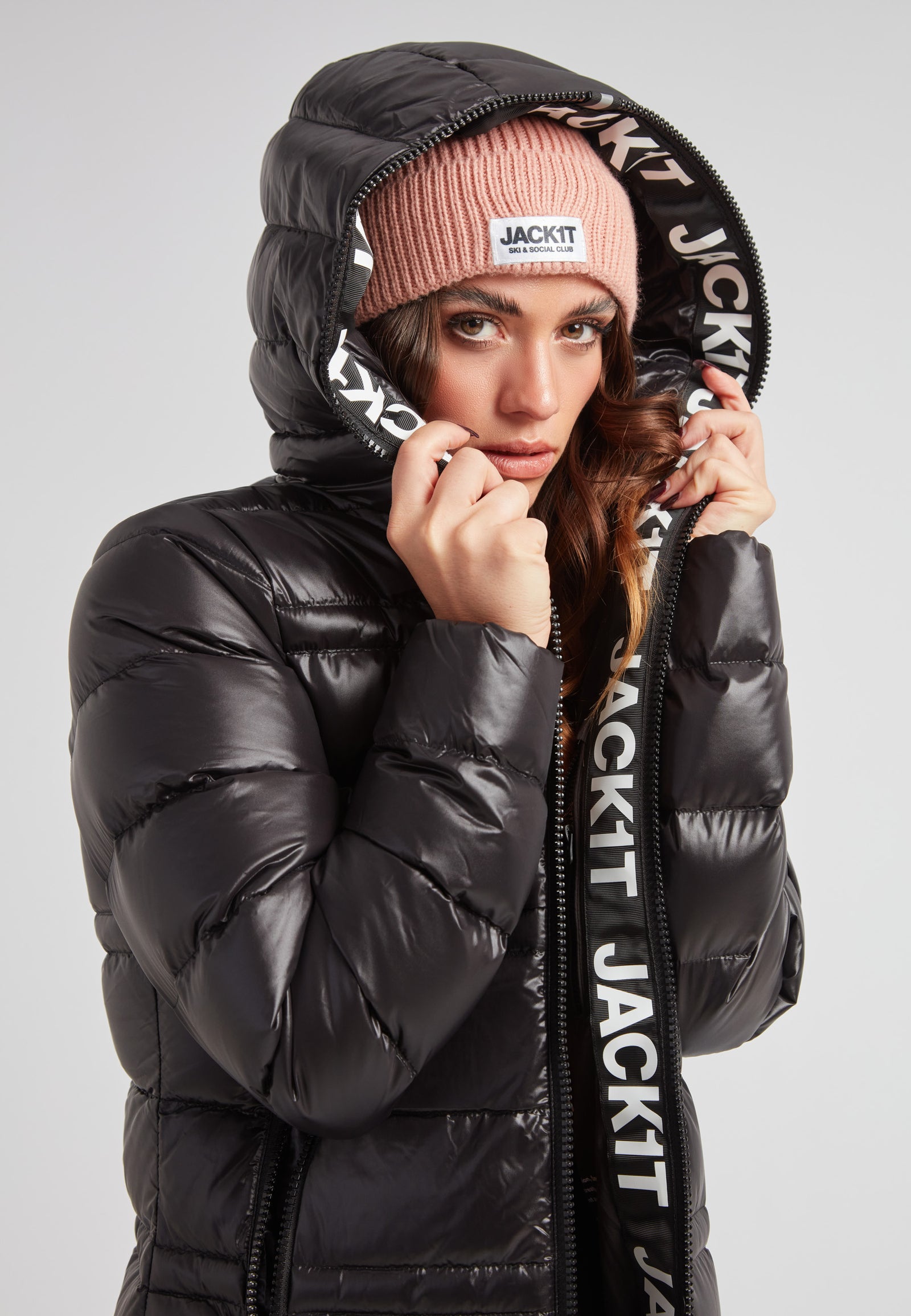 R4D Tramline Extra Long Down Coat Lux Black model wearing black coat with pink beanie, showcasing design and hood.