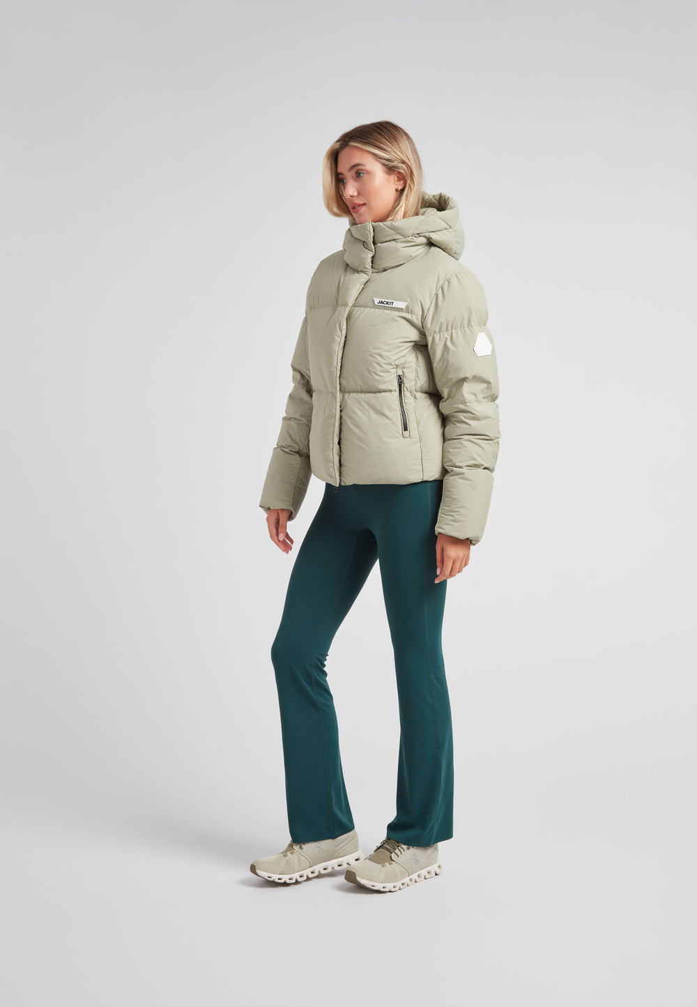 Woman wearing the Star Shower Down Puffer Jacket Sage, paired with green pants and stylish sneakers.